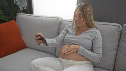 Wall Mural - Pregnant woman smiling while having a video call on smartphone at home, relaxing on a couch in a cozy living room, showcasing joyful anticipation of motherhood.