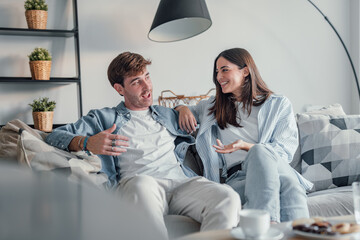 Young happy couple having fun talking laughing relaxing at home on couch, boyfriend embracing girlfriend telling funny joke sitting on sofa, humor in relationships, enjoying weekend together.