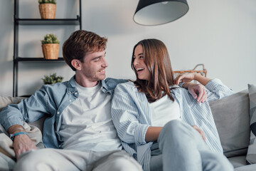 Young happy couple having fun talking laughing relaxing at home on couch, boyfriend embracing girlfriend telling funny joke sitting on sofa, humor in relationships, enjoying weekend together.