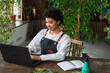 © Юлия Завалишина - African woman florist gardener working in botanical store using laptop order plants communicating on Internet with customer. Female small business owner working at flower shop. Running of own business