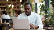 © elena - Serious millennial african-american man using laptop sitting at cafe table, focused black casual guy communicating online, writing emails, distantly working or studying on computer in public place