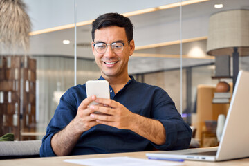 Aufkleber - Smiling busy professional Hispanic business man sitting in office holding mobile cellphone. Young happy businessman entrepreneur using smartphone looking at cell phone technology device at work.