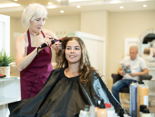 Wall Mural - Positive elderly female hairstylist using curling wands to create soft curls and add volume in long hair of young smiling female client..