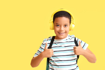 Wall Mural - Little African-American boy with school backpack and headphones showing thumbs up on yellow background