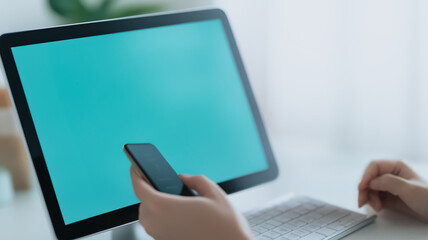 Poster - Hands Holding Phone Near Desktop: Close-up shot of hands holding a smartphone next to a desktop computer with a vibrant teal screen, suggesting seamless digital integration and modern workflow.