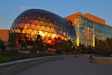 Poster - Modern Architectural Design in Sunset Light Featuring Reflective Facade and Creative Structure Surrounded by Lush Landscape and Vibrant Sky