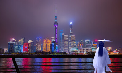 Wall Mural - A Chinese woman in traditional white dress holding a traditional white umbrella walks along - Shanghai colorful skyline with motion speed light of passing ferry in the foreground at night - Shanghai