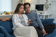 © crizzystudio - Relaxed asian couple sitting together on a comfortable sofa, enjoying a movie or tv show, changing channels with a remote control in their modern living room