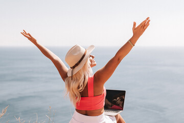 Wall Mural - A woman is sitting on a beach with her arms outstretched, wearing a pink top and a straw hat. She is looking out at the ocean, and there is a laptop on the ground in front of her.