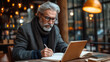 © Atchariya63 - An elderly man writes thoughtfully in a cozy cafe with warm lighting.
