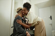 © Seventyfour - Side view of African American woman brimming with happiness giving gentle embrace to welcoming boyfriend in wheelchair with cute little French bulldog on lap at arrival home, copy space