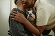 © Seventyfour - Portrait of Black woman gently hugging man while adorable French bulldog sitting on male owners lap and looking out with curious eyes indoors