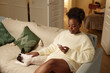 © Seventyfour - Medium shot of relaxed Black woman sitting on sofa with cute little dog, while looking at phone screen reading news feed or chatting cozy in living room