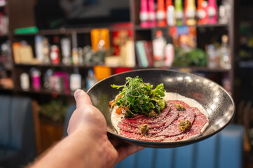 Wall Mural - A close-up of a hand holding a gourmet dish featuring thinly sliced meat, garnished with fresh greens and capers, presented in a stylish black plate against a blurred background of modern restaurant.