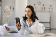 © fizkes - Mobile medical software. Happy woman doctor use telehealth app on phone at workplace manage electronic prescription communicate with healthcare team text message to patient calculate medication dosage