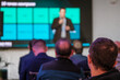 © Anton Gvozdikov - Blurry speaker in business presentation viewed over audience head. Professional event atmosphere, networking focus.