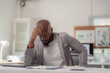 © Tj - Tired african american entrepreneur suffering from headache while reviewing financial reports, sitting at workplace with laptop and documents, feeling exhausted and stressed from overwork