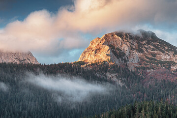 Wall Mural - Beautiful sunrise illuminating a rocky mountain peak with a cloudy sky and fog covering the forest