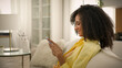 © Yuliia - Smiling African American girl woman play mobile video game on home couch in living room happy relaxed ethnic female teenager scrolling smartphone social networks addict internet dating apps side view