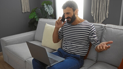 Wall Mural - Handsome young hispanic man with a beard talking on the phone while sitting on a couch in a living room, working on a laptop.