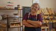 © Krakenimages.com - Hispanic woman with grey hair and glasses, arms crossed, standing in a bakery filled with bread and pastries, smiling confidently.