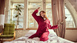 © master1305 - Smiling, happy young woman in red pajamas sitting in bed with white top, cheerfully dancing a home in warmly lit bedroom. Concept of happiness, carefree lifestyle, relaxation