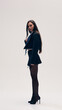 © master1305 - Full-length side view image of young beautiful woman posing in cropped jacket and shirt with a tie and heeled shoes against studio background. Concept of business, fashion trends, beauty, style