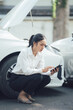 © Kritdanai - Roadside Assistance: A young businesswoman crouches beside her broken-down car, using her phone to call for help.  The scene is calm, yet conveys a sense of urgency and the need for assistance.