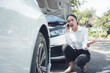 © Kritdanai - Distressed Woman Calling for Roadside Assistance: A young woman in business attire crouches beside her broken-down car, urgently making a phone call for roadside assistance.