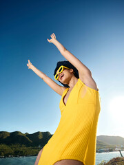 Poster - Woman in a yellow dress lifting her arms, wearing a sun hat against a clear blue sky