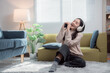© Tj - Happy young woman sitting on the floor and singing using her smartphone as a microphone, enjoying music with headphones in her modern and cozy apartment