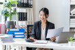 © Kritdanai - Focused Businesswoman at Desk: A smiling, young businesswoman sits at her desk, reviewing paperwork and using a calculator, showcasing efficiency and professionalism in a modern office setting.