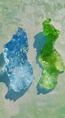 Poster - Two colorful ice cubes, one blue and one green, melting on a textured green surface, casting distinct shadows that highlight their form and the melting process