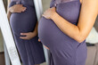 © Vadim - Expecting mother admiring her baby bump. A pregnant woman in a polka dot dress softly touches her belly while gazing into a mirror at home.