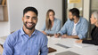 © fizkes - Young 20s Arabian male employee posing for camera seated at desk with colleagues on background, exudes competence, professionalism and reliability. Bank advisor, financial consultant portrait. Career