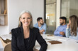 © fizkes - Experienced professional mature woman with gray hair and confident expression sitting at table in modern conference room with teammates on background. Executive manager, financial consultant portrait