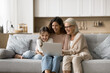 © fizkes - Funny movie. Friendly family of 3 generation mid aged granny young mother cute little tween age daughter grandchild spend free time hugging on sofa using notebook make shopping online watch video blog