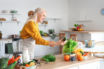 Wall Mural - Mature woman with fresh vegetables and fruits making healthy smoothie in kitchen