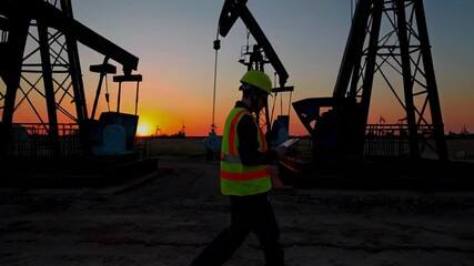 Leinwandbilder - Engineer in protective gear walking through oil field at sunset, carefully inspecting pump jacks using tablet while analyzing operational data and monitoring industrial extraction processes