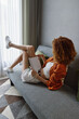 © SHOTPRIME STUDIO - Stylish young woman relaxing on a couch, reading a book, wearing casual summer attire with curly hair, creating a cozy and vibrant atmosphere