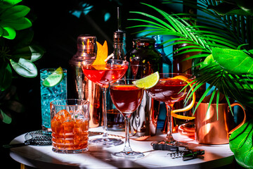 Wall Mural - Summer alcoholic cocktail party with ready-made cocktails, bar tools and bottles of spirits on marble table with tropical plants. Green background, hard light and shadows. Shallow DOF