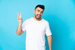 © luismolinero - Caucasian man over isolated blue background smiling and showing victory sign