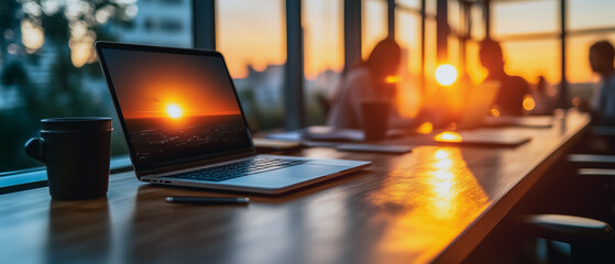 Poster - Sunset Office Vibes: Laptop displaying a vibrant sunset on its screen sits on a wooden table in a modern office, with blurred silhouettes of colleagues in the background.