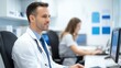 © Tondone - A focused male doctor working at a computer in a modern medical office, emphasizing professionalism and dedication to patient care alongside a female colleague in the background.