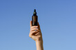 © Daryna  - Female hand holding brown glass jar of serum with pipette dispenser on blue sky background. Concept of beauty and aesthetics products, skin care, hydration and nutrition