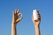 © Daryna  - Female hand holding white capsule pill and white mockup jar of pills on blue sky background. Concept of pharmacy, medicine and natural dietary supplements for health support and treatment