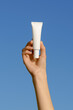 © Daryna  - Vertical image of a female hand holding a white mockup tube of cream on a blue sky background. Concept of facial skin care, cosmetology, beauty products, serum, aesthetics