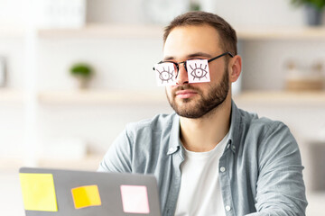 Poster - Tired millennial guy having stickers with drawings of open eyes on his glasses, sleeping in front of laptop at home office. Exhausted young man overworking, falling asleep a workplace