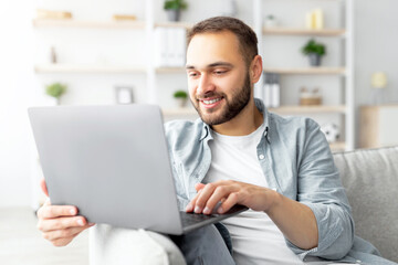 Poster - Cheerful millennial man sitting on couch with laptop computer, working or studying online from home office. Joyful Caucasian guy participating in web lecture, taking part in video conference