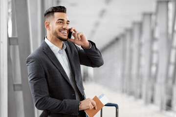 Wall Mural - Handsome Middle Eastern Businessman Talking On Cellphone While Waiting In Airport Lounge, Successful Arab Entrepreneur Standing With Luggage Suitcase In Terminal And Enjoying Mobile Call, Copy Space